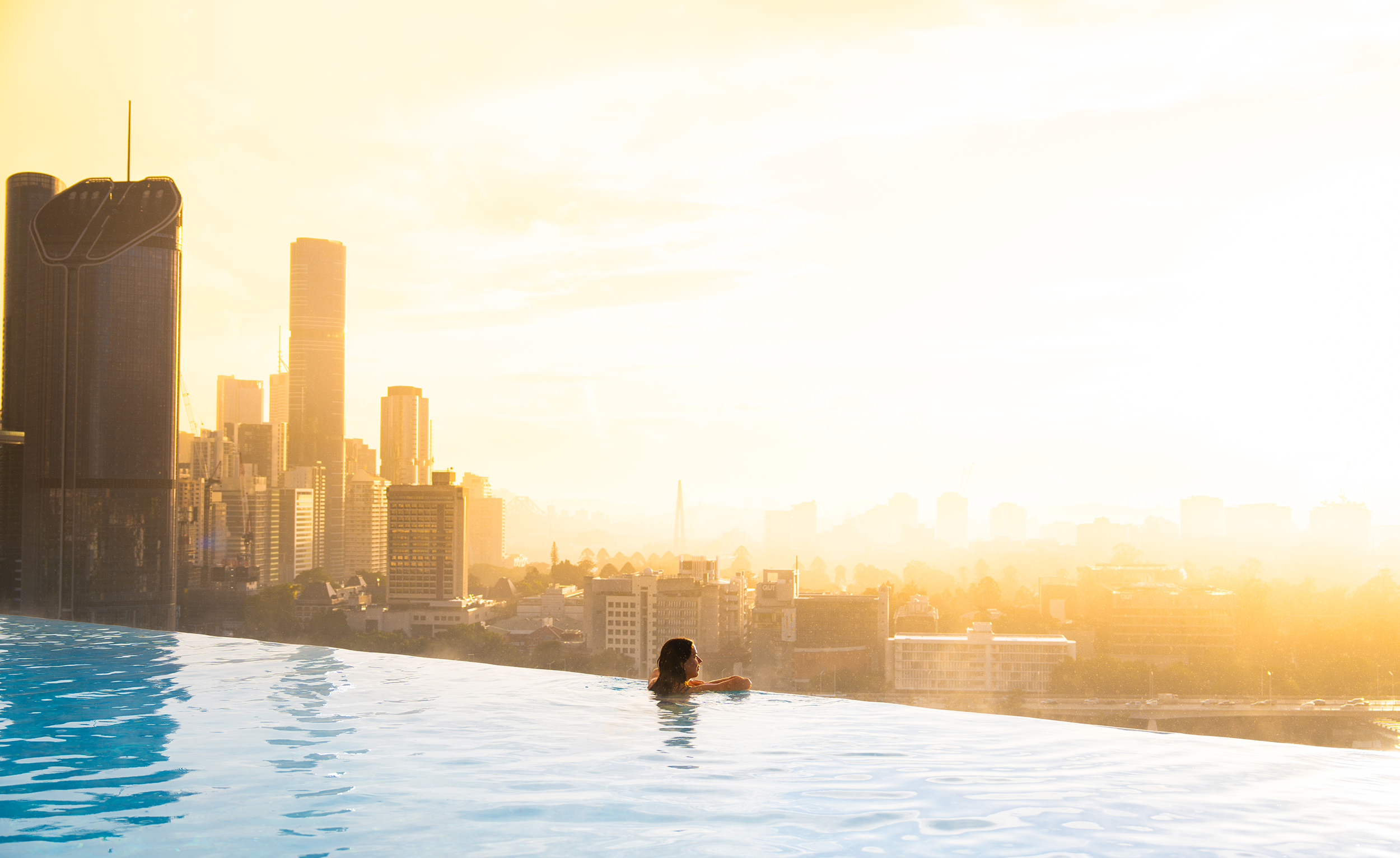 A person swims in an infinity pool overlooking a city skyline at sunset, with tall buildings and a warm golden glow filling the scene.