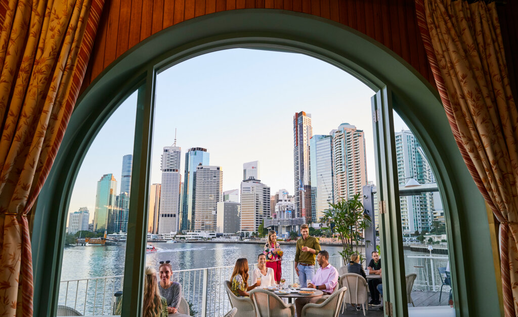 People dining on a terrace by a river, with tall city buildings in the background, viewed through a large arched window with curtains.