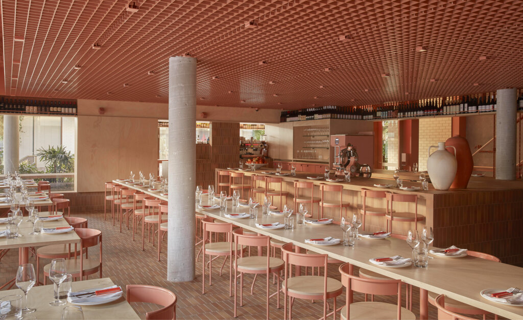 A modern restaurant with peach-colored decor, wooden tables set with glassware, pink chairs, and a bar area. Shelves of wine bottles line the upper walls. Pillars and plants are visible inside.