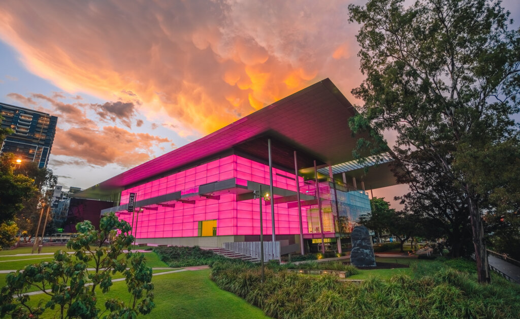 A modern building with glowing pink walls stands in a park at sunset, with dramatic orange clouds overhead and trees surrounding the structure.