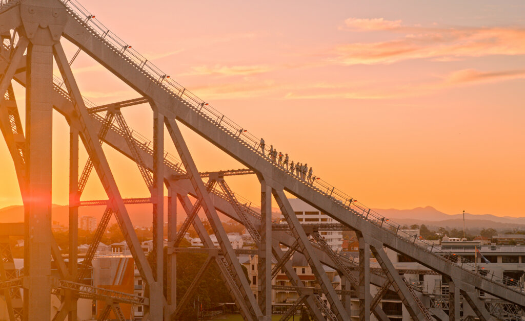 People climb a steel bridge structure at sunset, with a warm orange sky, city buildings below, and distant mountains in the background.