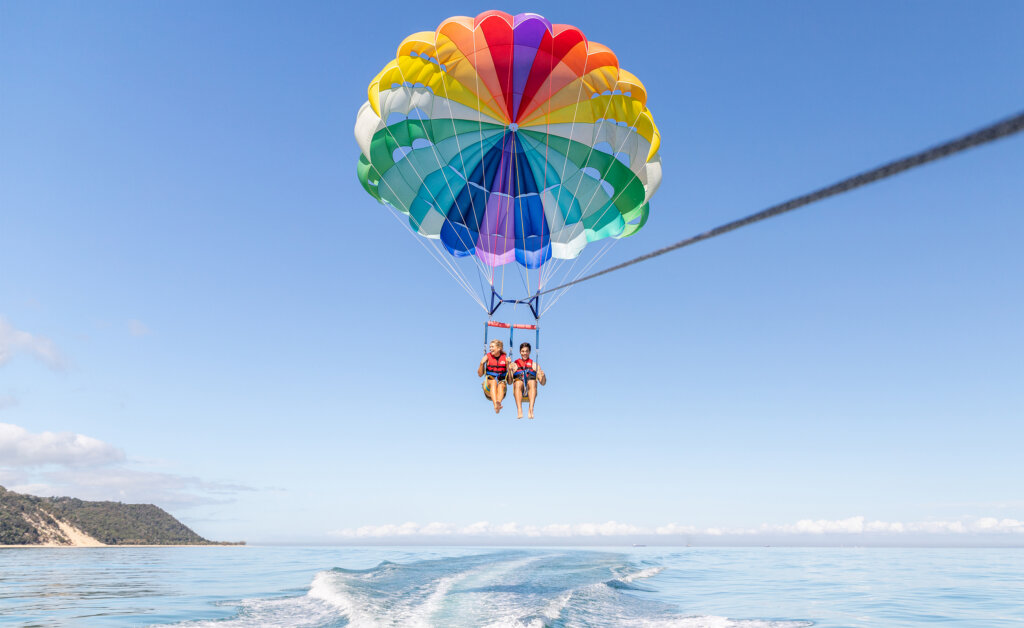 Two people parasailing over the ocean, suspended by a colorful parachute, with a boat creating waves below them and a distant shoreline visible on the left.