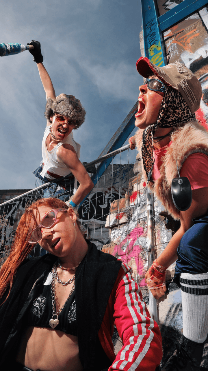 Three people in edgy, punk-inspired outfits pose energetically in front of a graffiti-covered wall and fence, with dramatic facial expressions and dynamic body language against a blue sky.