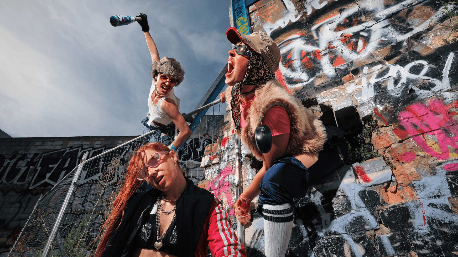Three people in eccentric, punk outfits pose energetically by a graffiti-covered wall; one shouts, another raises a bottle, and the third leans forward wearing sunglasses. The scene is vibrant and rebellious.