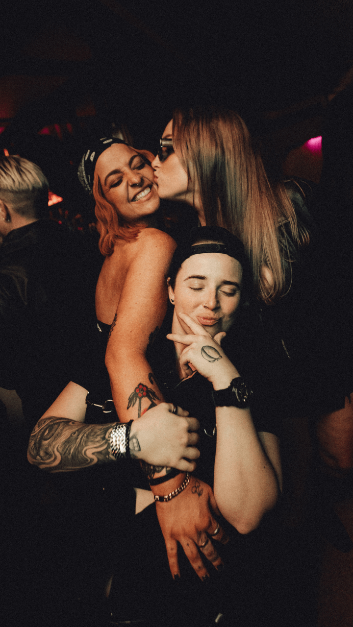 Three friends pose together at a party; one person kisses another on the cheek while the third smiles and poses with a peace sign. The group appears happy and relaxed in a dimly lit setting.