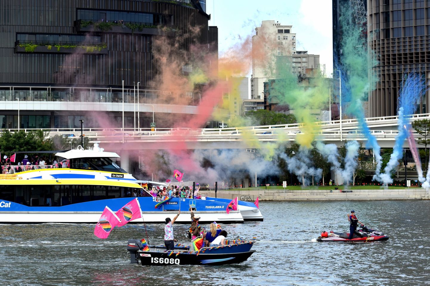People on boats celebrate on a river with colorful smoke and flags. A large ferry and modern buildings are in the background. The sky is partly cloudy.