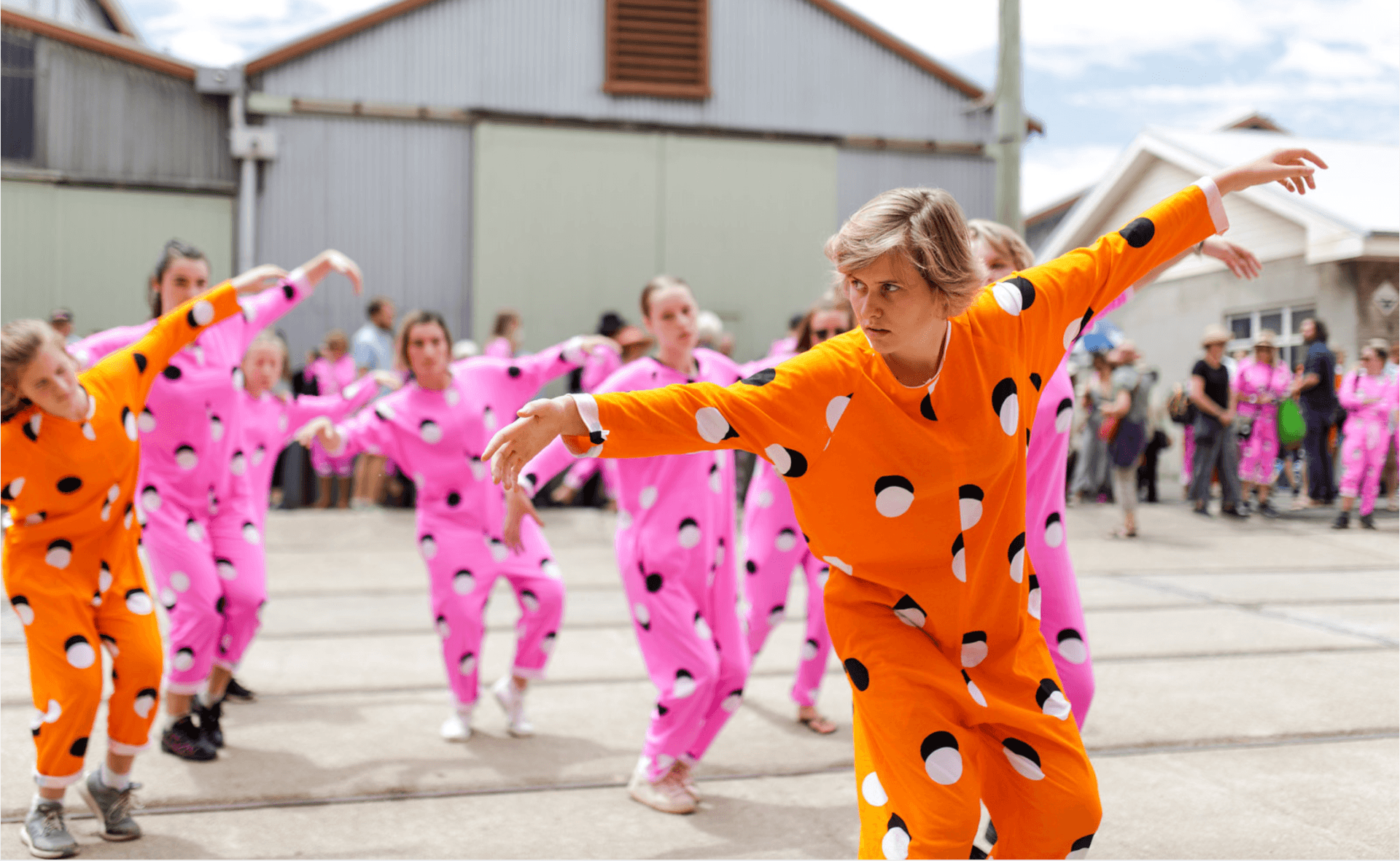 A group of people in pink and orange polka-dot costumes dance outdoors on concrete, with focused expressions. Industrial buildings and a crowd are visible in the background.