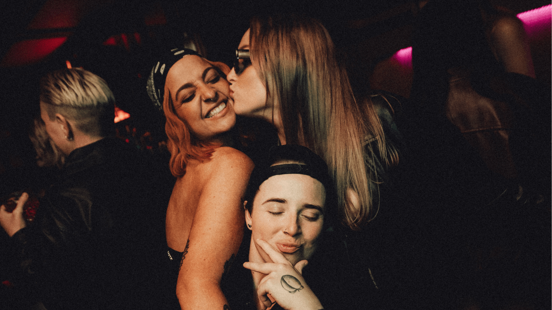 Three people pose together at a dimly lit party or nightclub. One person kisses another on the cheek, while the third person in front smiles with eyes closed, making a peace sign gesture.