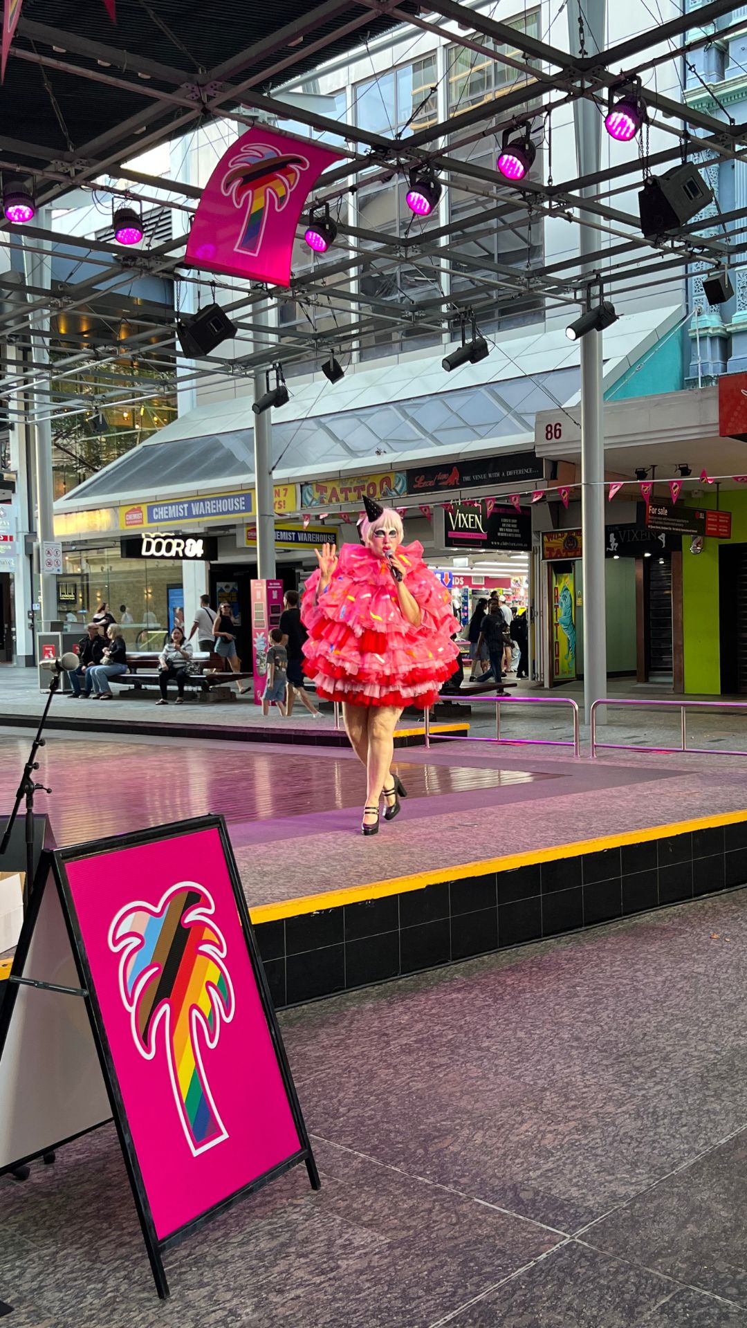 Performer in a pink ruffled costume dances on an outdoor stage beneath colorful lights. A sign with a palm tree logo is in the foreground; shops and people are in the background.