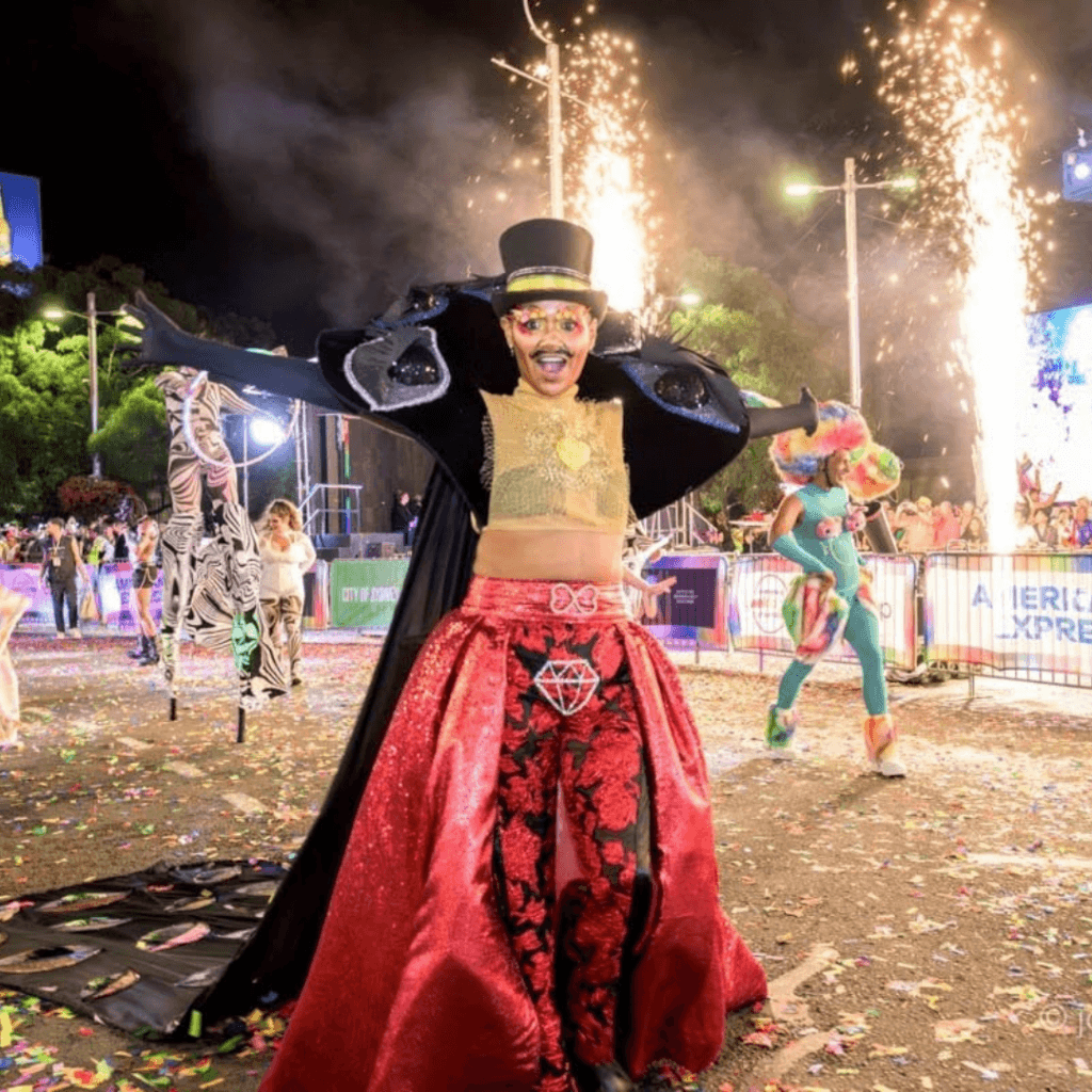 A performer in a top hat, gold crop top, and red skirt with a diamond emblem dances in the street, flanked by pyrotechnics and other performers.