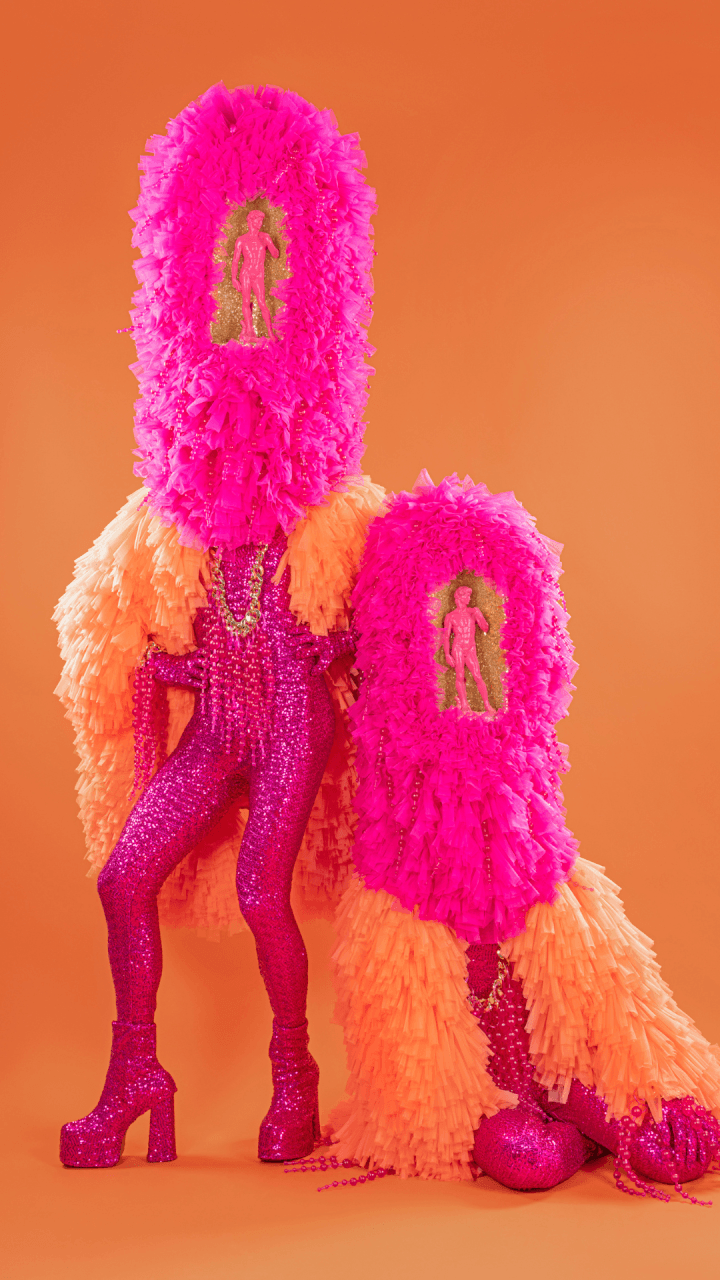 Two people in bright pink and orange costumes with tall, fringed headpieces featuring cutouts displaying small pink figurines, posing against an orange background.