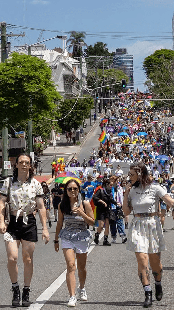 A large crowd marches up a city street during a Pride parade, with people holding rainbow flags and wearing colorful outfits under a sunny sky. Four individuals walk in the foreground.