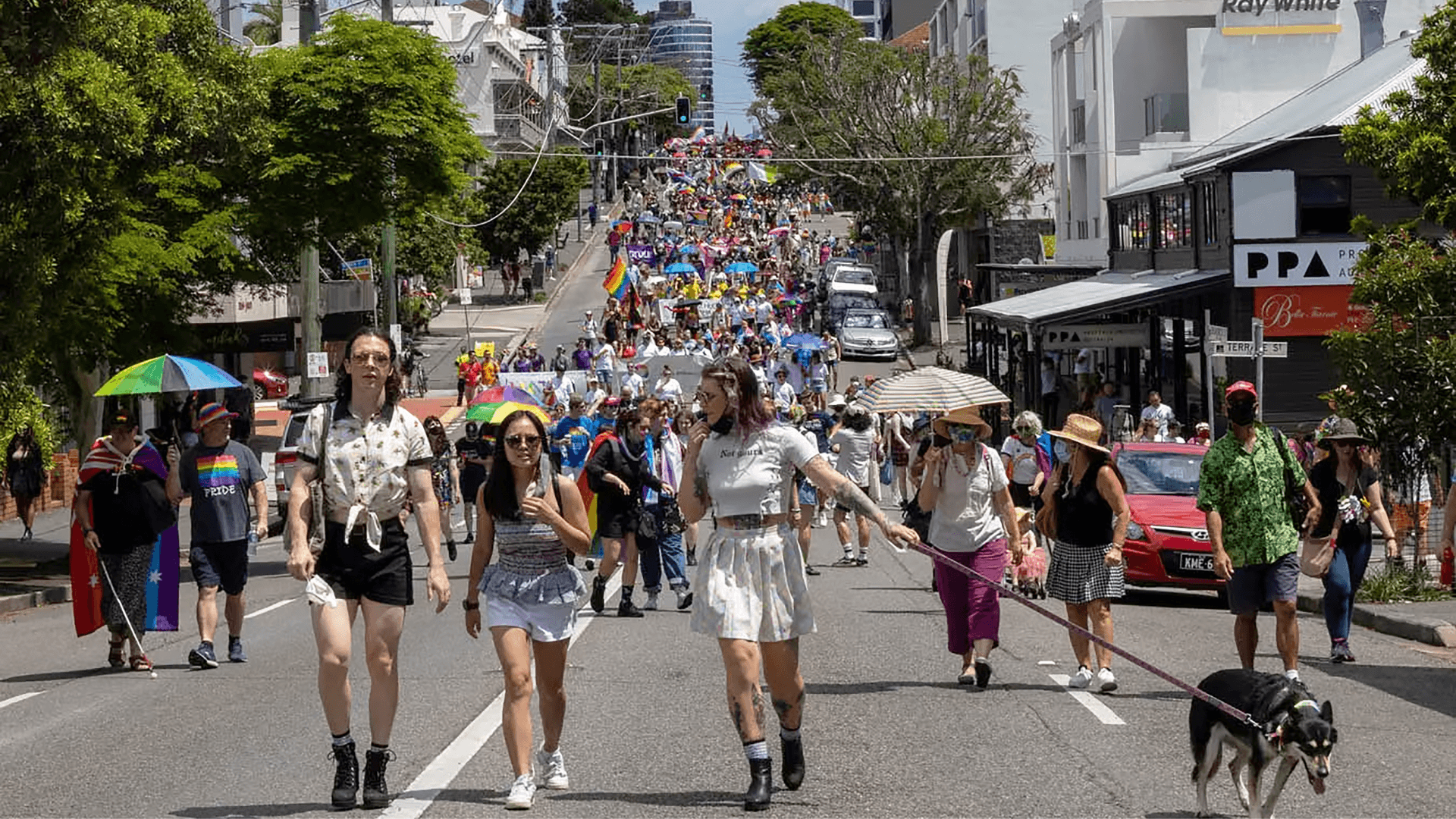 A large crowd marches up a city street during a pride parade, carrying rainbow flags. People in colorful outfits walk in front, some with dogs, and others use umbrellas for shade on a sunny day.