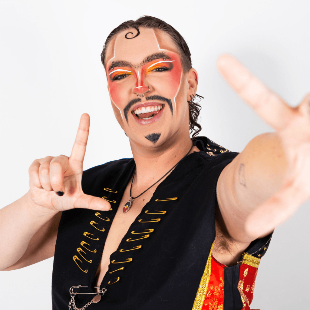 A smiling person with bold orange and red face paint, a mustache, and a black vest adorned with gold safety pins, making a finger frame gesture toward the camera.