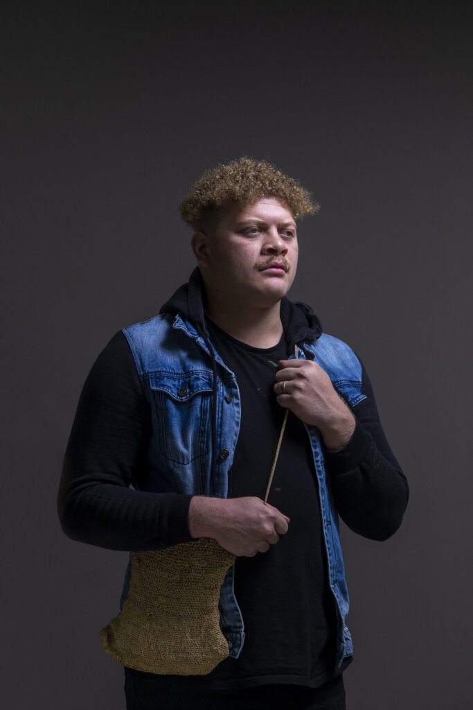A person with curly hair wears a denim vest over a black hoodie, holding a woven bag and a thin stick, standing against a dark gray background while looking slightly to the side.