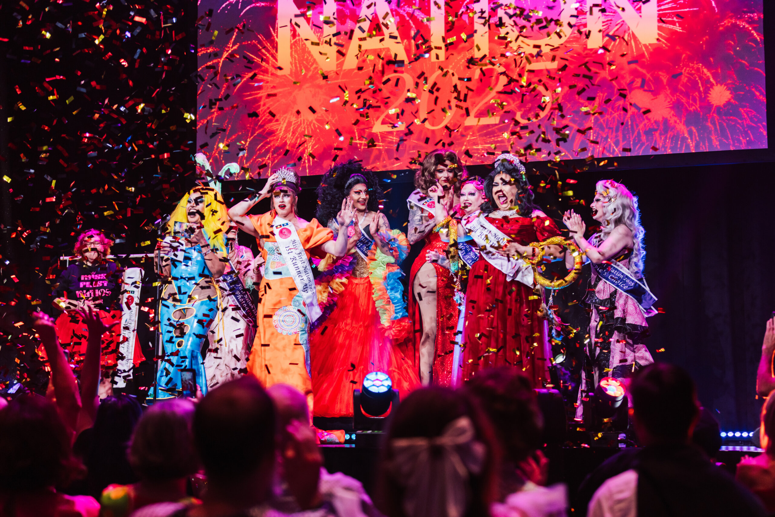 A group of drag performers in colorful gowns celebrate on stage under falling confetti, with an audience cheering. A large illuminated sign reading NATION 2023 glows in the background.