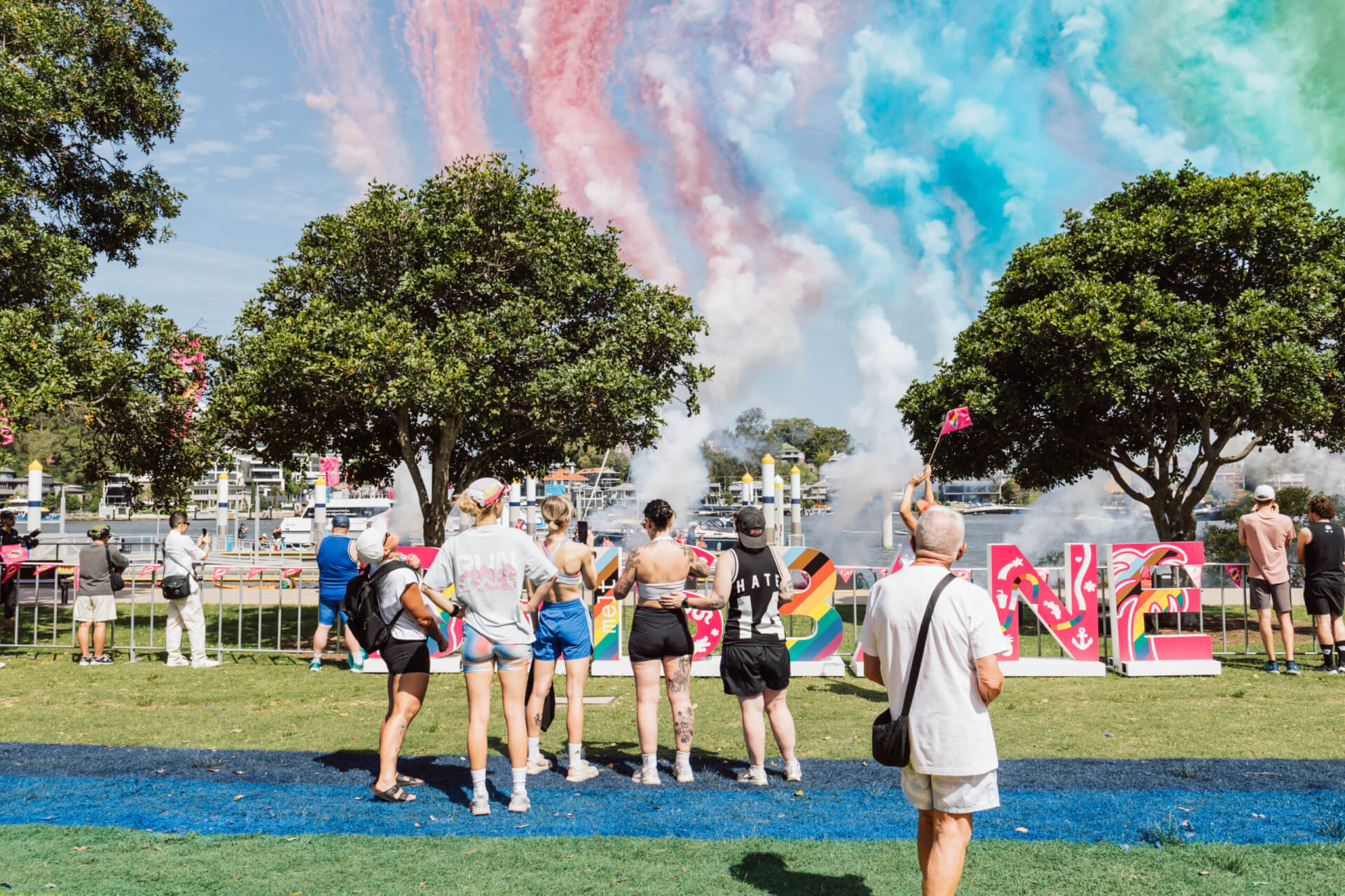 People stand on grass watching colorful smoke trails in the sky at an outdoor event. Large decorated letters and trees are visible, with water and boats in the background.