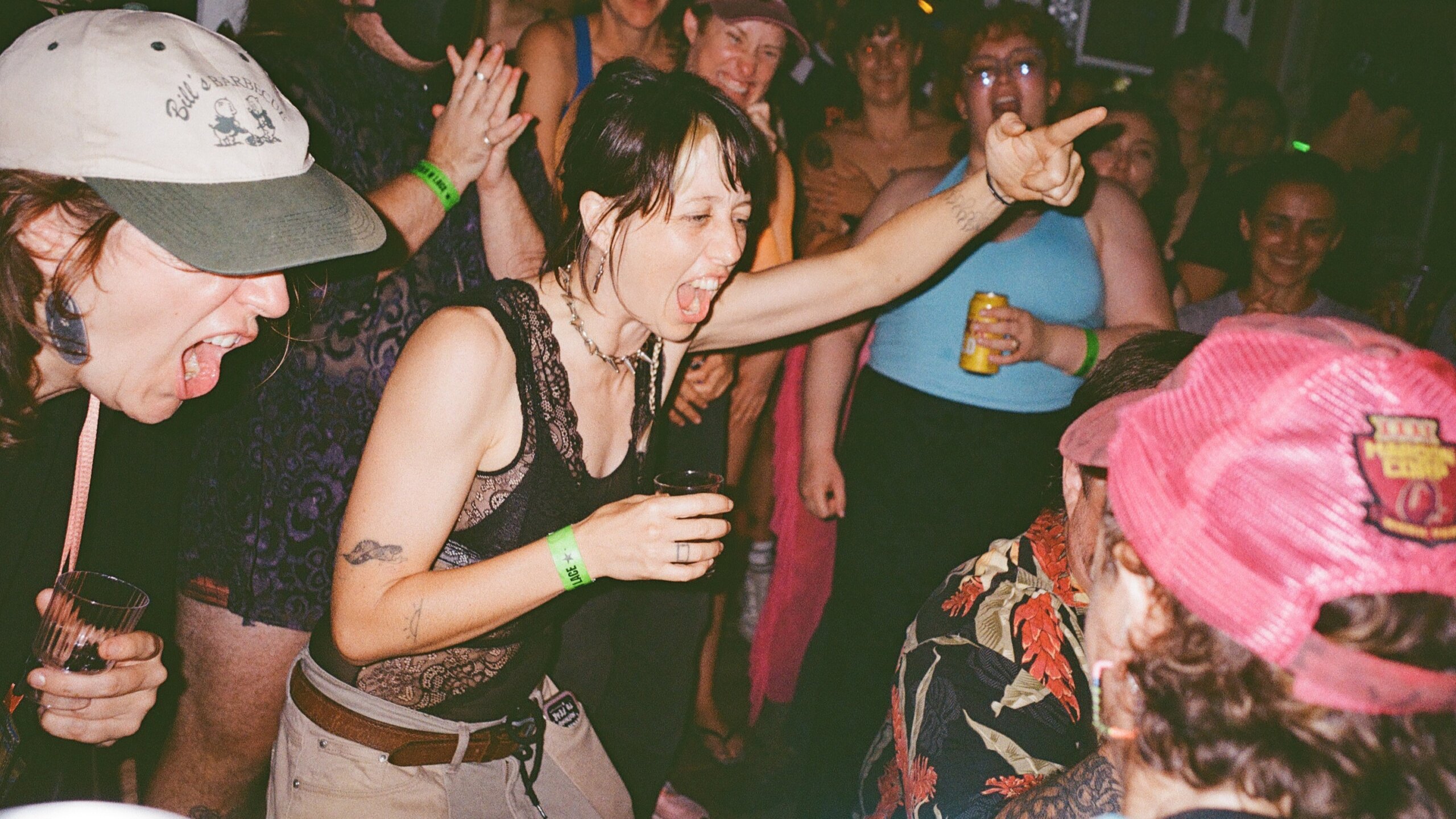 A woman shouts with her arm outstretched in a lively, crowded room. People around her cheer and smile, some holding drinks, creating an energetic party atmosphere.