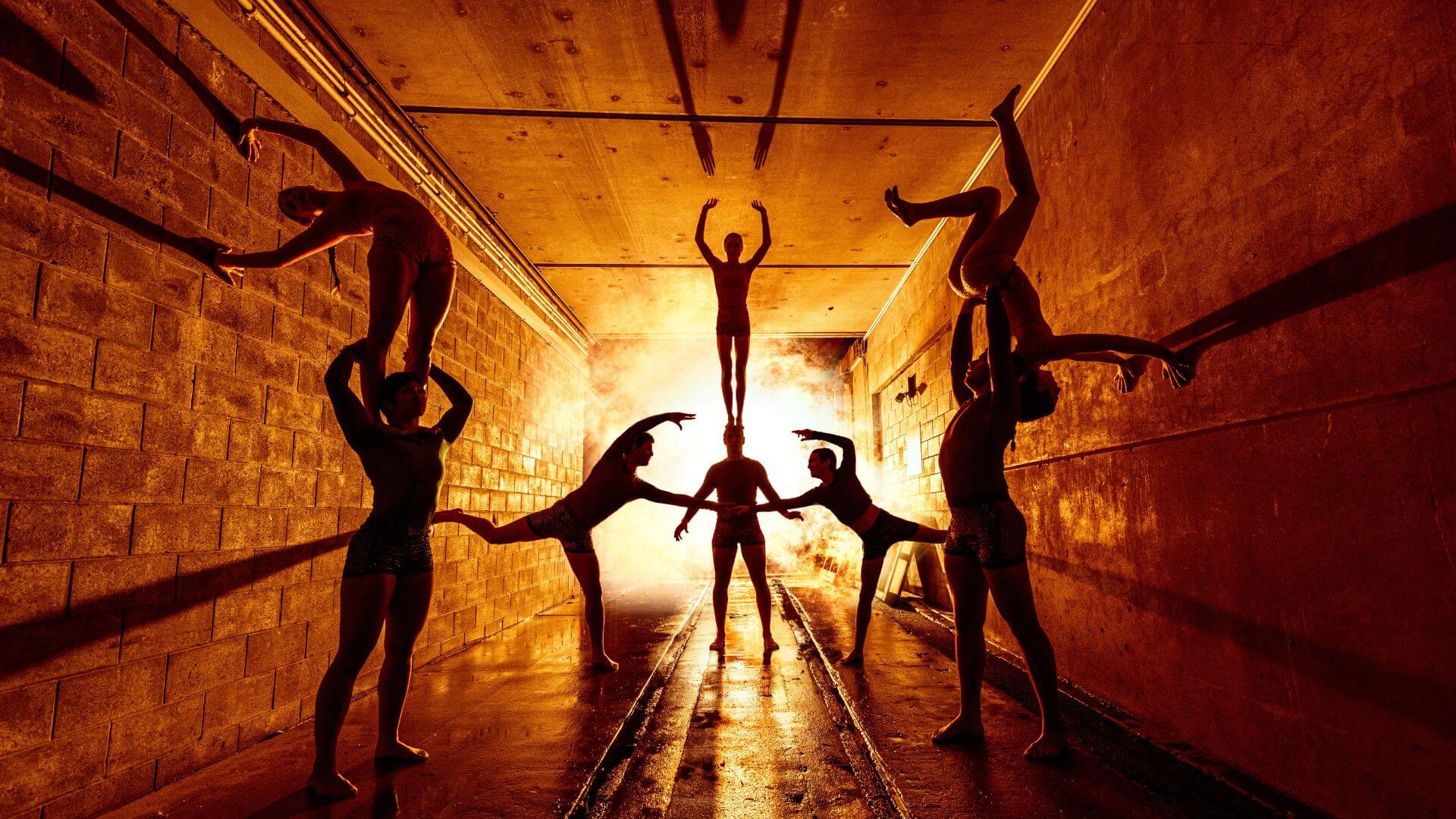 Seven acrobats perform balancing acts in silhouette inside a tunnel, with dramatic orange light and flames illuminating the scene from the background.