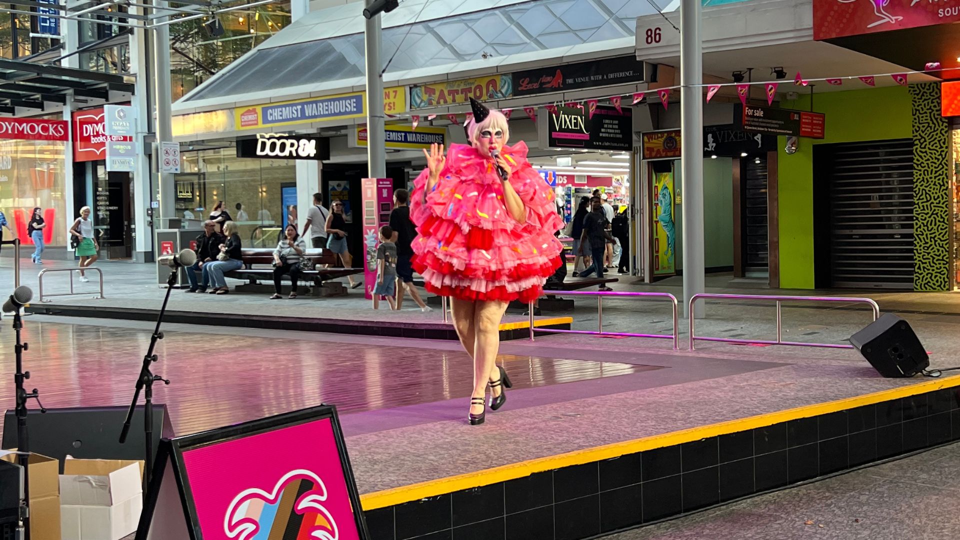 A performer in a bright pink ruffled dress and high heels walks on an outdoor stage in a shopping area. People watch in the background. Pink decorations and storefronts are visible.