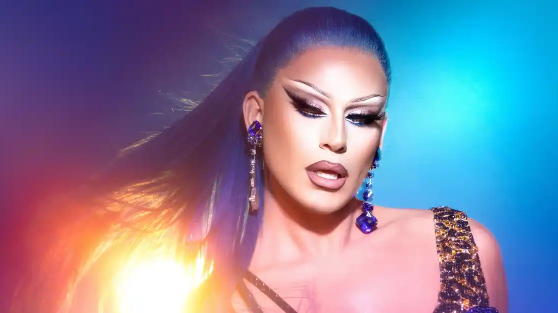 A person with long, sleek blue hair and dramatic makeup poses in front of a vibrant blue and orange light. They wear sparkling blue earrings and a sequined outfit, looking confidently toward the camera.