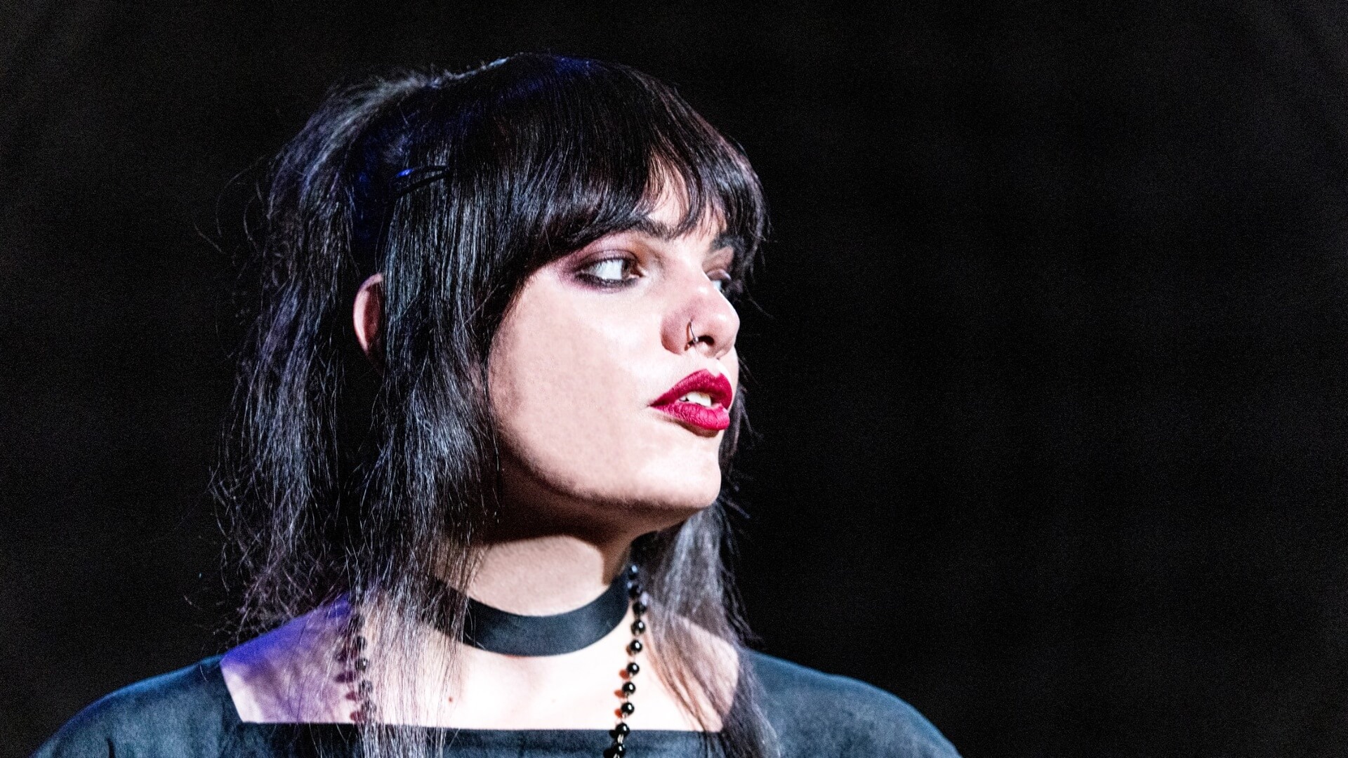 A person with long dark hair, bold red lipstick, and dark eye makeup gazes to the side. They wear a black choker and a dark top, with a blurred dark background.