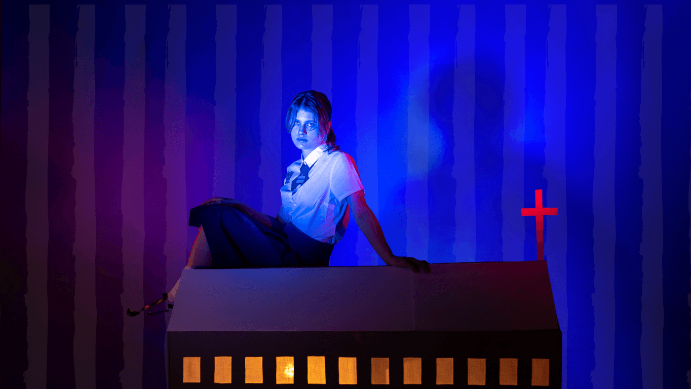 A woman in a school uniform sits on a model church roof, illuminated by blue and red light, with yellow-lit windows and a glowing cross on the right against a striped background.