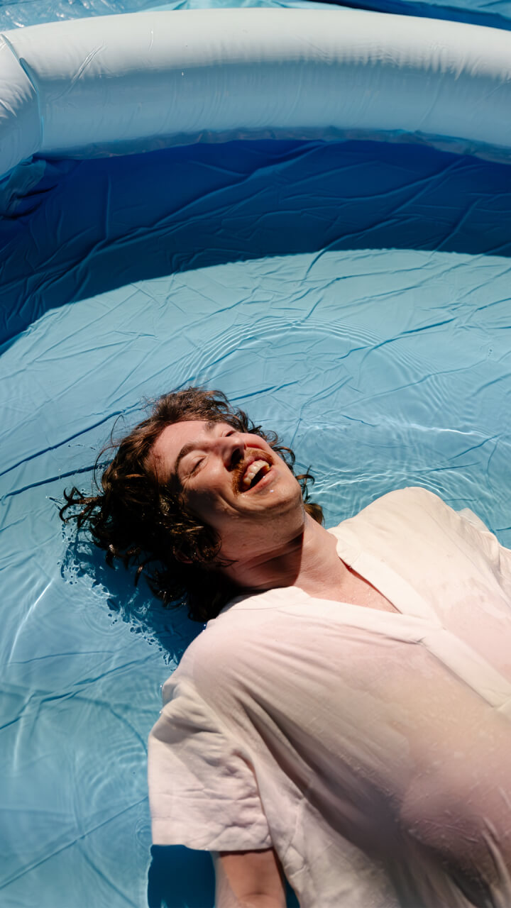 A person with curly hair and a mustache lies on their back, smiling and relaxed, in a shallow inflatable pool filled with water, wearing a light-colored shirt.