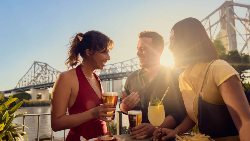 Three people stand by a riverside table with drinks and food, laughing and talking at sunset. A bridge is visible in the background, and sunlight shines between them, creating a warm, lively atmosphere.
