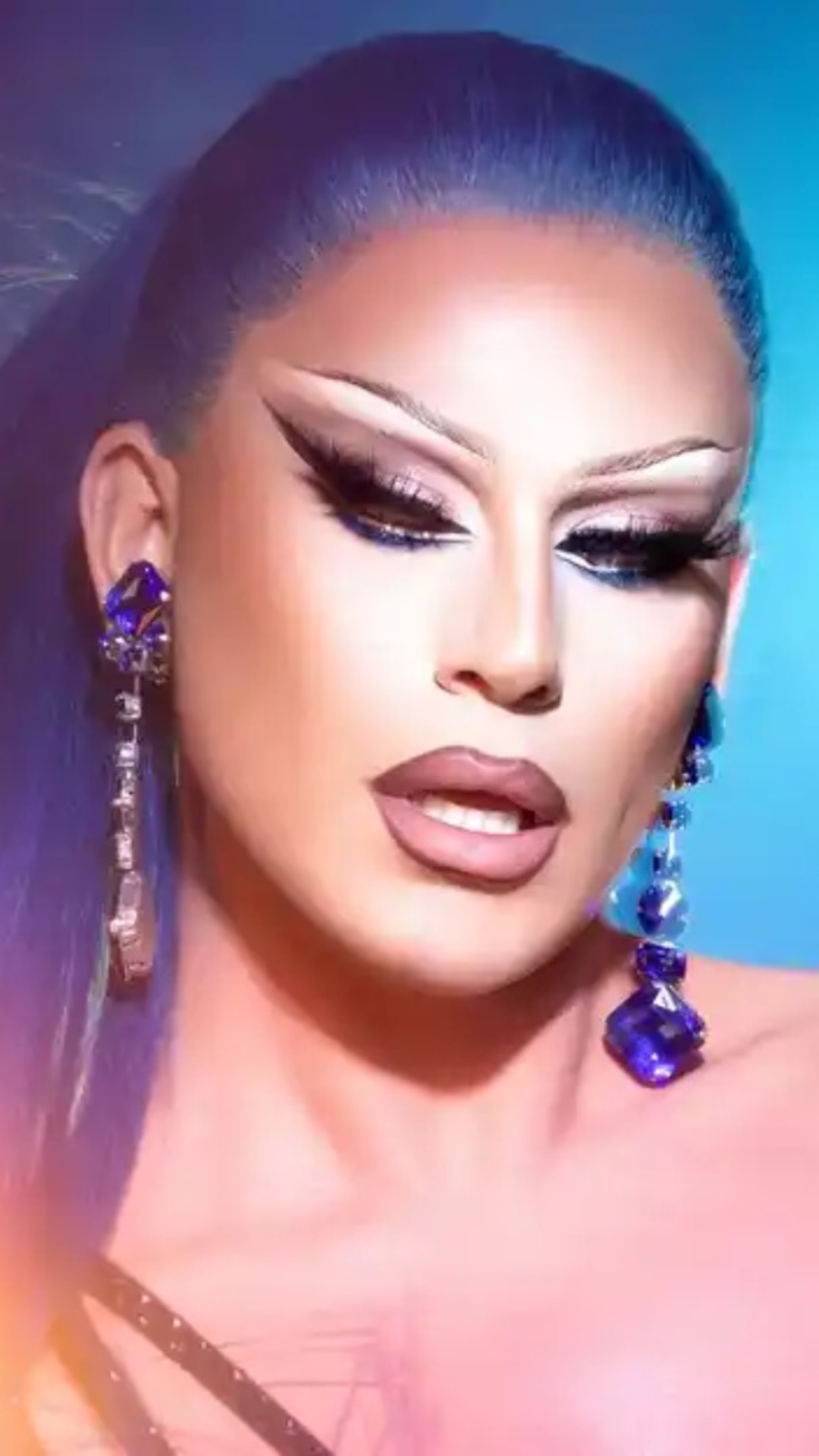 Joey Jay with long blue hair, dramatic winged eyeliner, bold eye makeup, nude lips, and large dangling blue gemstone earrings against a blue background.