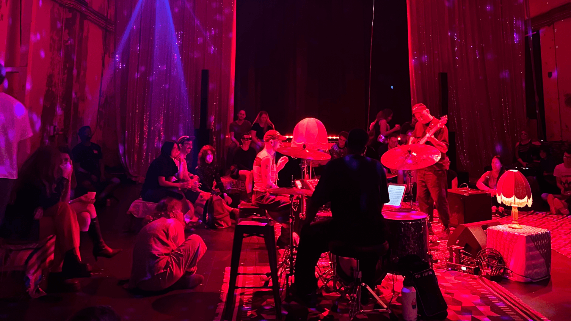 A band performs on stage under red lighting as an audience sits closely around them, some on the floor. Sparkling curtains and lamps add to the intimate, cozy atmosphere.