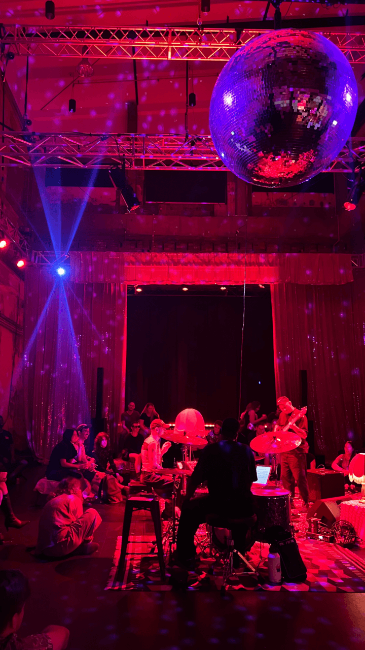 A band performs on a dimly lit stage with red lighting, surrounded by an audience sitting on the floor. A large disco ball hangs above, casting blue and red reflections across the room.