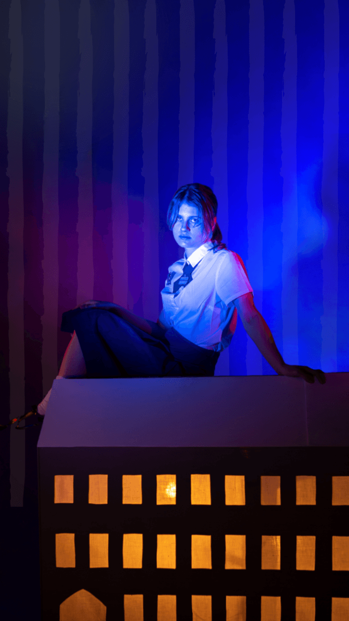A person in a white shirt and dark pants sits on a lit model house, illuminated by blue and pink lights, with vertical striped walls behind them.