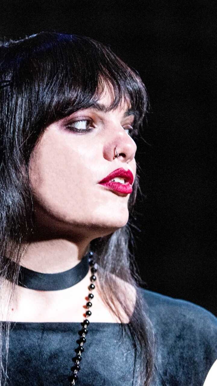A person with long dark hair, bold red lipstick, a nose ring, and dramatic eye makeup looks to the side. They wear a black choker and beaded necklace against a dark background.