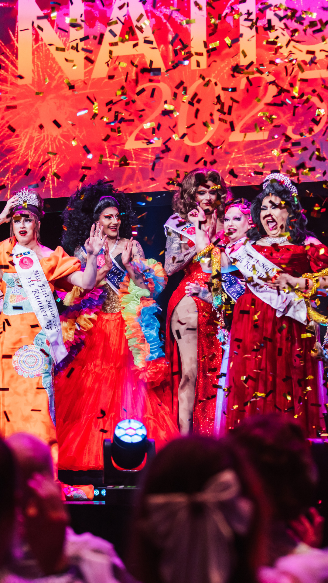 Drag performers in colorful gowns celebrate on stage under confetti, wearing crowns and sashes, with a bright screen displaying 2023 and fireworks in the background.
