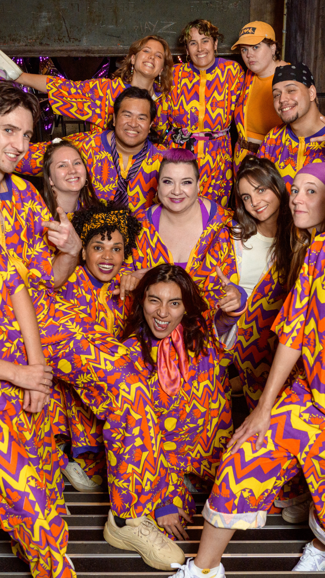 A group of people pose together, most wearing vibrant orange, purple, and yellow zigzag-patterned outfits, while two wear plain clothes and hats. They smile and appear joyful, standing on steps with festive decorations in the background.