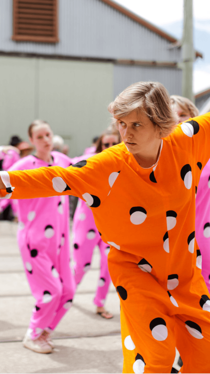 A person in an orange, polka-dotted costume stretches their arms out while others in matching pink costumes dance in the background outdoors.