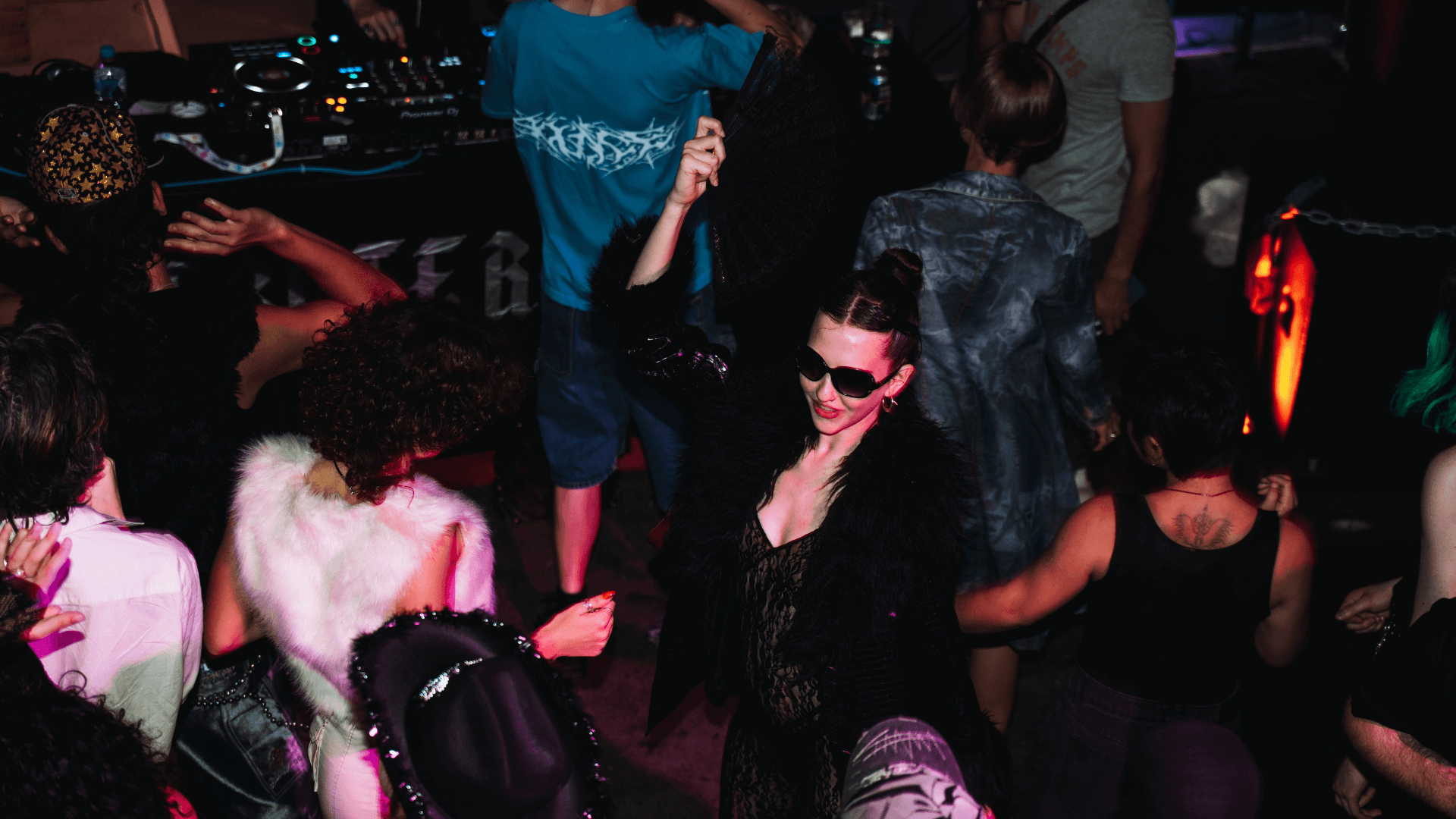 A group of people dance at a lively indoor party; one woman in the center, wearing sunglasses, black clothes, and holding a fan, stands out among the crowd.