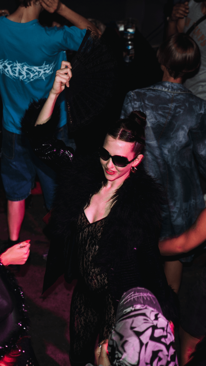 Woman in sunglasses and black outfit dances at a party, holding a black fan above her head. Other people in casual, colorful clothing dance around her in a dimly lit setting.