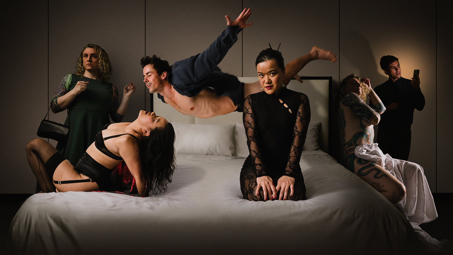 Six people pose dramatically on and around a bed in a dimly lit room; each has a distinctive outfit, expression, and pose, creating a surreal and theatrical scene.