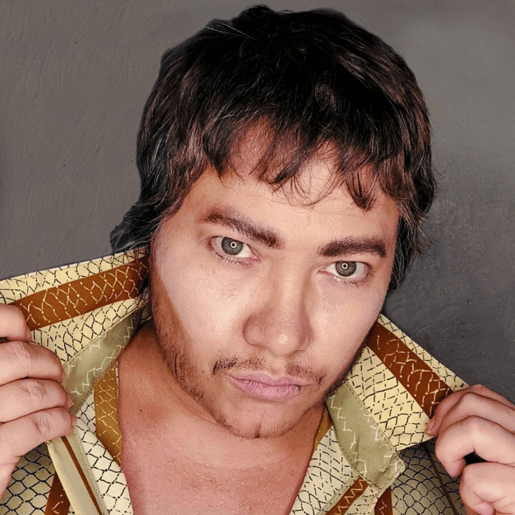 A person with light brown hair and facial hair holds the collar of a patterned tan and brown collared shirt.