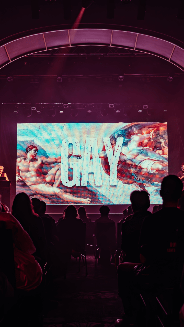 Audience watches a stage presentation featuring a large screen displaying Michelangelos art, The Creation of Adam, with the word GAY in bold white letters across the center.