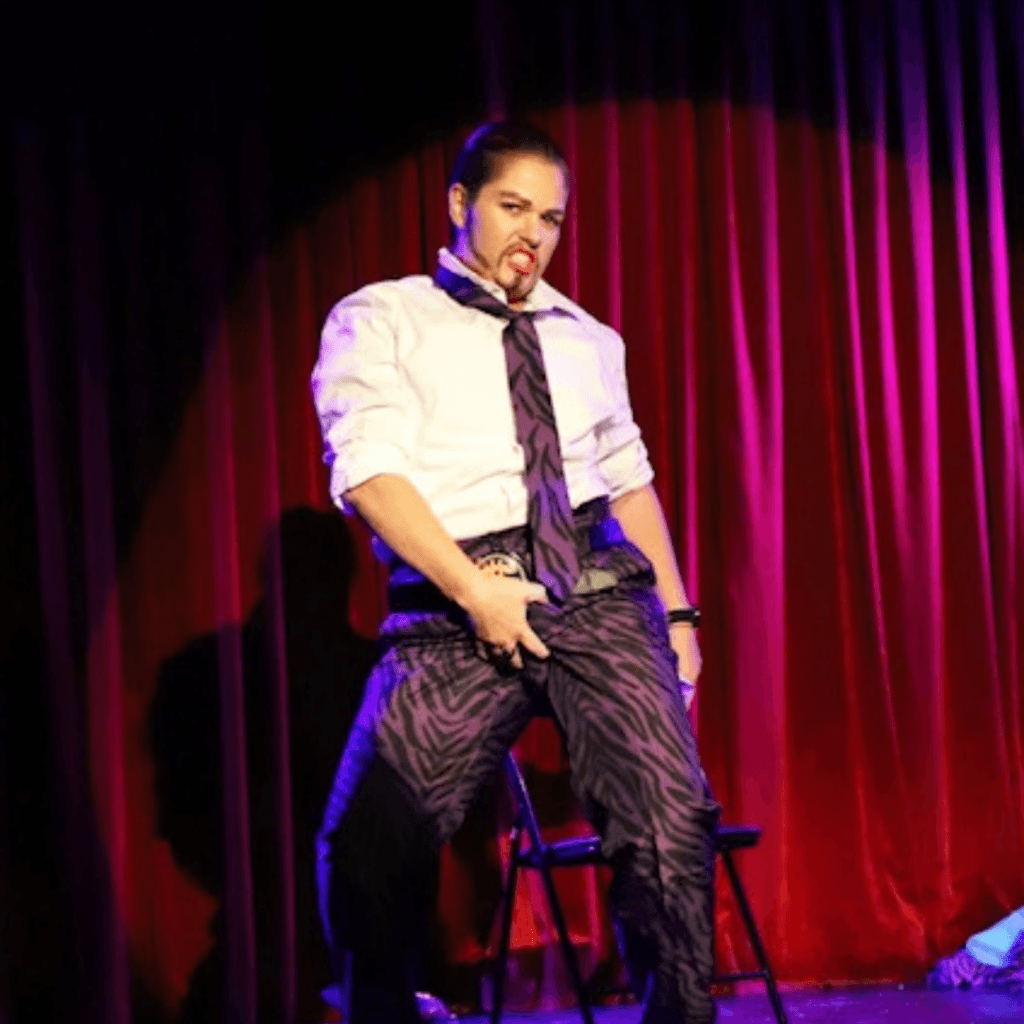 A performer in a white dress shirt and purple animal-print tie and trousers poses suggestively in front of red stage curtains.
