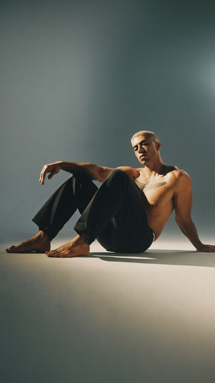 A shirtless person with short blond hair sits on the floor with one knee raised, wearing black pants, in soft lighting against a neutral background.