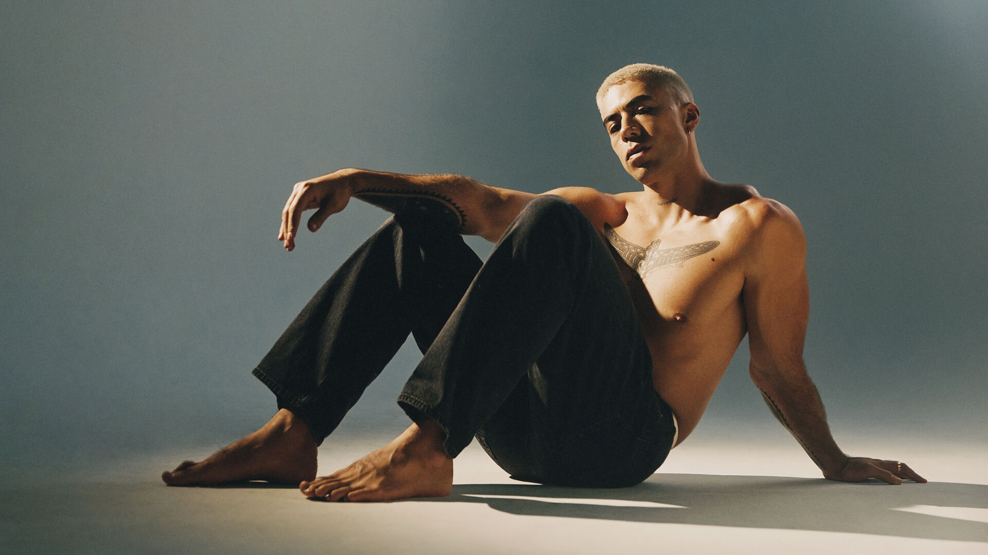 Shirtless person with short blond hair and dark jeans sits on the floor in a studio setting, looking relaxed with one knee up and an arm resting on it. Soft, warm lighting creates subtle shadows on the background.