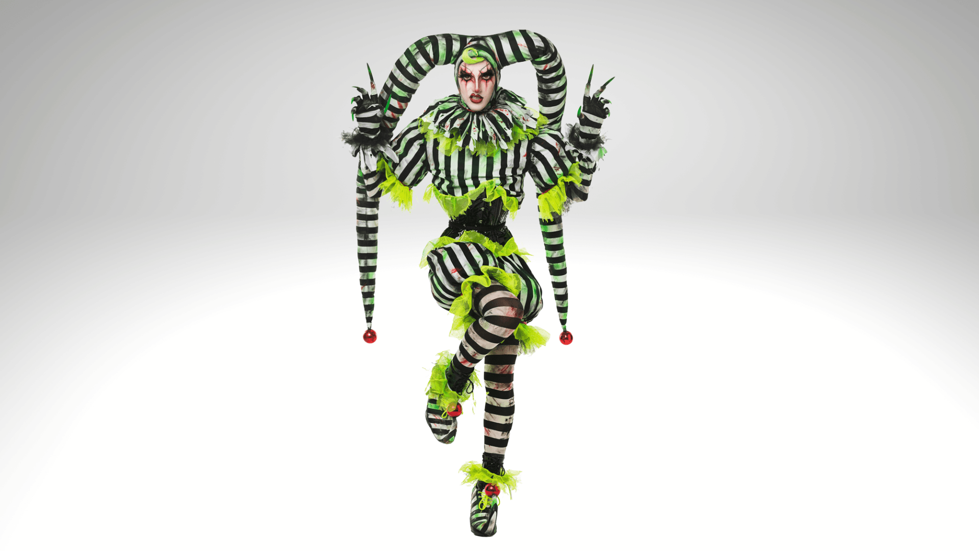 A person poses in a jester costume with black and white stripes, neon green accents, and two long horn-like hats. They stand on one leg with arms raised, and their face is painted white with dramatic makeup.