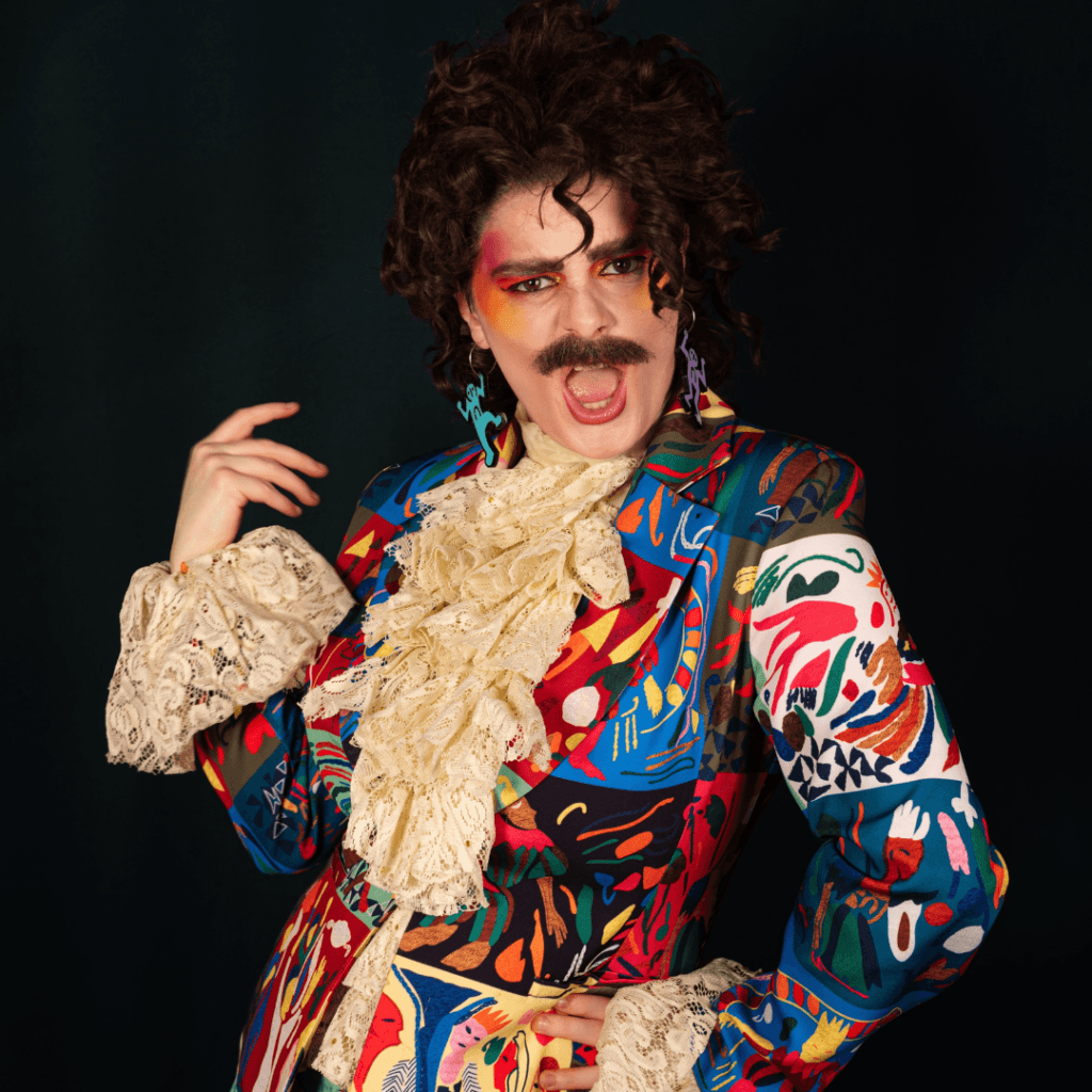 A person with a mustache and curly dark hair wears a vibrant, multicolored patterned blazer with a lace jabot and gecko-shaped earrings.