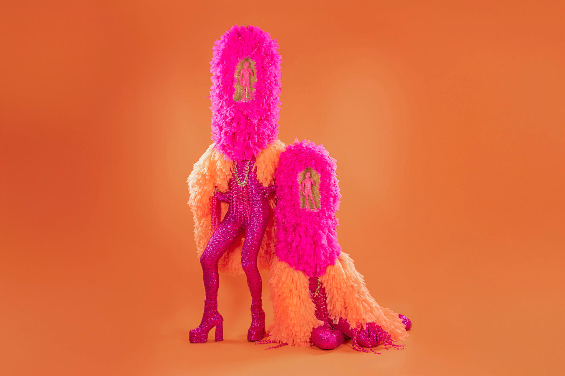 Two figures in bright pink and orange textured costumes with tall headpieces stand against an orange background. Their faces are covered, and their outfits have sparkling details and platform boots.