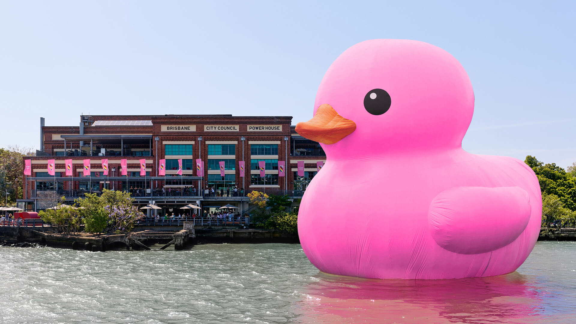 A giant pink rubber duck floats on a river in front of a building labeled Brisbane City Council Powerhouse under a clear sky.