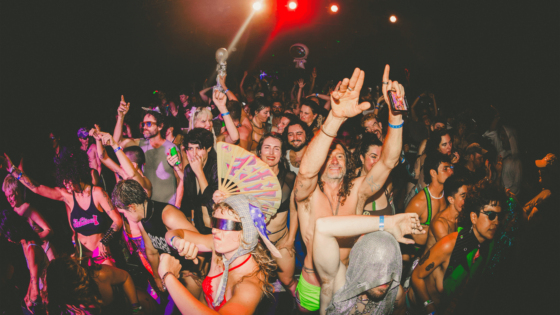 A crowd of people dancing and celebrating at a vibrant party, many wearing colorful, eccentric outfits and accessories, with bright stage lights shining above them.